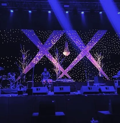A starry night theme for a Sangeet stage, using a backdrop of fairy lights, crisscross drapes, and a central chandelier to create a magical setting for the live band.
