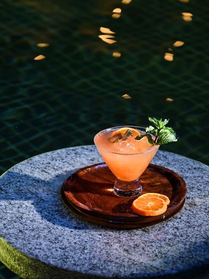 A vibrant orange cocktail rests on a stone table by the pool, its surface reflecting the gentle evening light. Garnished with fresh mint, it’s the perfect companion for a tranquil moment.