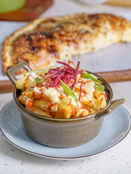 A bowl of loaded fries with a cheesy calzone in the background. This shot captures a full, hearty meal, perfect for a casual restaurant or cafe menu. The focus is on the toppings and textures of the fries.