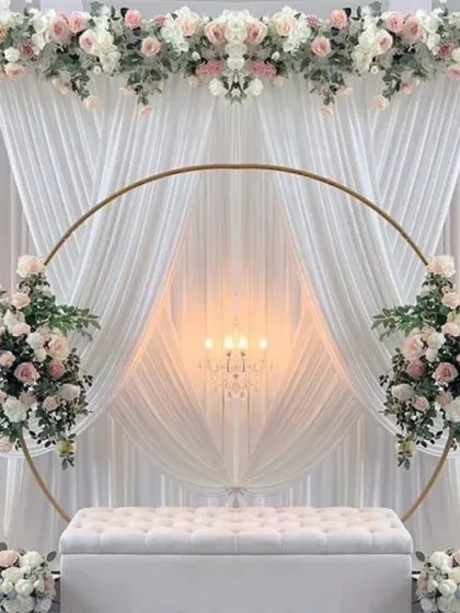 An absolutely breathtaking wedding or engagement stage. The setup features a large golden ring, elegant white draping, and beautiful arrangements of white and pink artificial flowers.