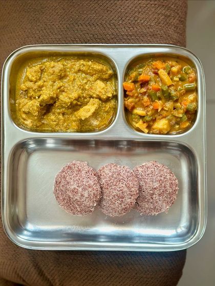 This South Indian thali features red rice idlis with chicken curry and a side of mixed vegetable curry.