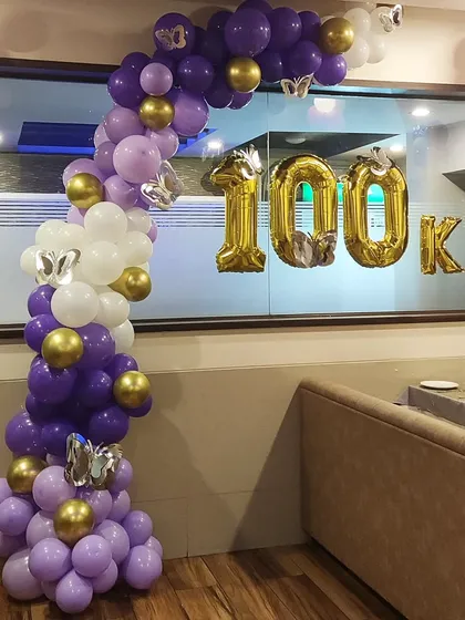 Celebrating a major milestone with a custom balloon arrangement. This organic half-arch in purple, white, and gold is paired with large '100K' foil balloons to mark the achievement, perfect for social media announcements or company events.