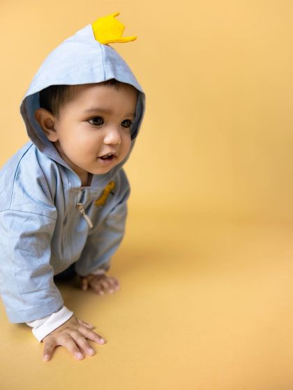 A curious toddler in a blue dinosaur-themed hoodie crawls towards the camera on a bright yellow studio background. This action shot captures the exploratory nature of early childhood.
