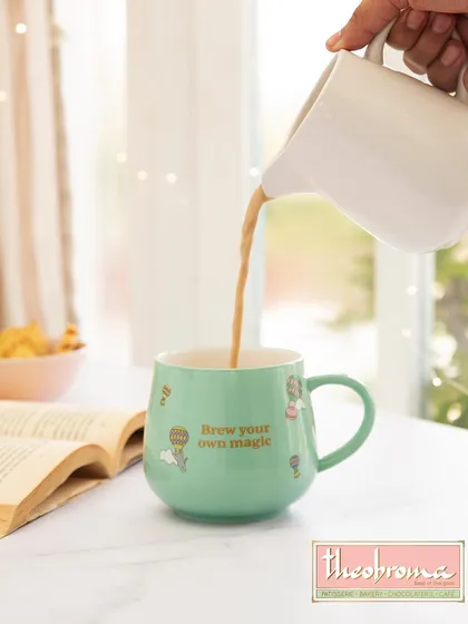 A lifestyle product shot for Theobroma, capturing a quiet moment of indulgence. The action of pouring tea into the branded mug, set against a soft, book-filled background, creates a cozy and aspirational scene.