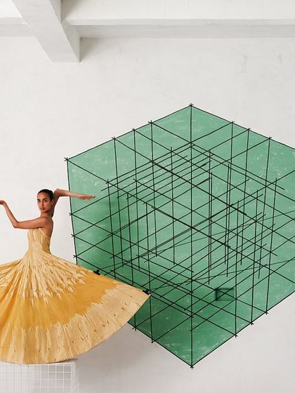 A model in a yellow Banarasi drape appears to float in front of a green geometric installation, creating a surreal and colorful composition.