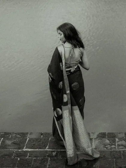 A full-length back profile in black and white. This artistic shot focuses on the silhouette and the beautiful drape of the saree against a minimalist background, emphasizing form and texture.
