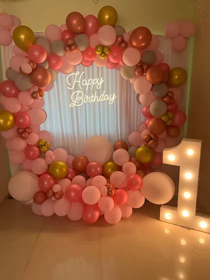 An elegant circular balloon arch in shades of pink and rose gold for a first birthday. A large light-up marquee number '1' stands beside it, creating a perfect photo spot.