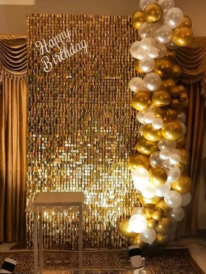 An elegant birthday photo corner at home. A gold sequin wall provides the shimmer, complemented by a cascade of white and gold balloons and a simple 'Happy Birthday' sign.