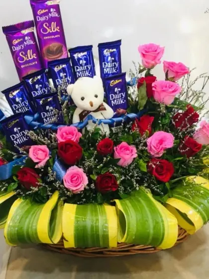 This basket arrangement is a perfect gift for someone special, combining flowers, chocolates, and a cute teddy bear. It features pink and red roses with a variety of Dairy Milk chocolates, creating a full and thoughtful hamper.