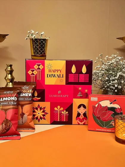 A close-up of a festive hamper, showcasing the colorful packaging, healthy snacks, and a decorative candle holder, all set against a traditional backdrop with brass diyas.