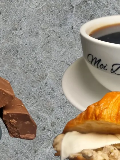 A perfect cafe moment. This image captures a cup of black coffee alongside a hearty croissant sandwich and a side of chocolate, showcasing a classic pairing.