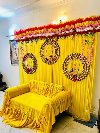 For a home mehndi or haldi function, I can create a vibrant photo corner like this one. This setup includes a bright yellow backdrop with matching sofa covers, decorative dreamcatcher-style hangings, and a floral border on top.