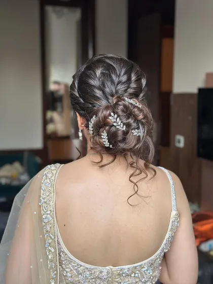 This messy bun features interwoven braids and is decorated with delicate silver leaf accessories. The loose curls around the face add a touch of softness, making it ideal for an engagement or reception.
