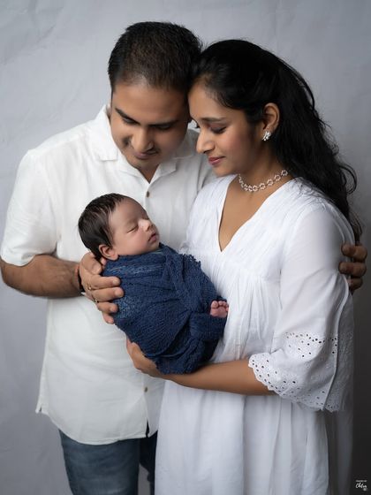 A classic family pose, with both parents holding their precious newborn. The coordinated white outfits create a clean and timeless aesthetic against the simple studio backdrop.