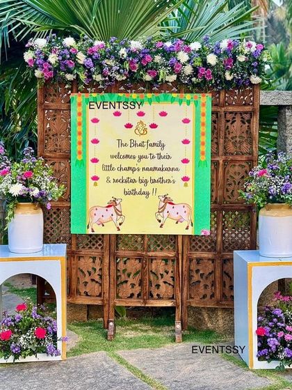 The welcome signage for the naming ceremony, set against a traditional carved wooden screen and flanked by floral arrangements. This entrance piece introduces the theme and welcomes guests to the sacred event.