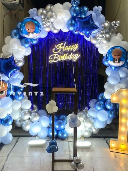This balloon arch decoration for a Boss Baby first birthday uses a blue foil curtain as the backdrop. The arch is made of blue, white, and silver chrome balloons, accented with star-shaped foils and a 'Happy Birthday' neon sign.
