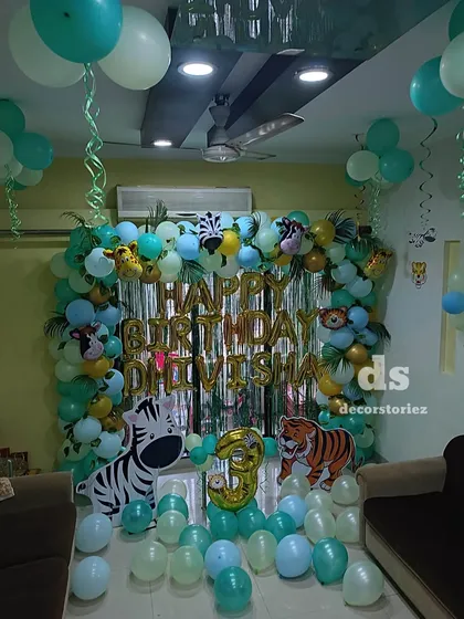 A jungle safari theme for a third birthday, featuring a lush balloon garland in shades of green and gold, a shimmer backdrop, and large zebra and tiger cutouts.