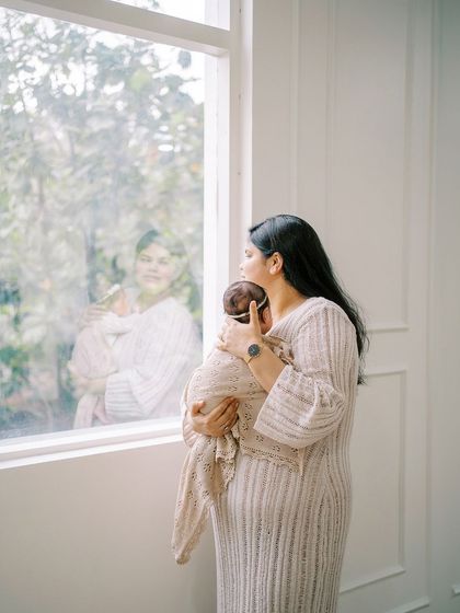 A mother holding her swaddled newborn by the window, with her reflection creating a beautifully layered and thoughtful portrait.