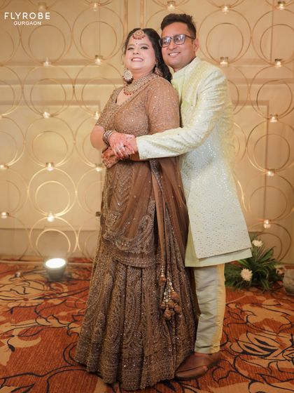 A happy couple posing for a photo at a wedding function. He is wearing a light green sherwani that perfectly complements her brown lehenga.
