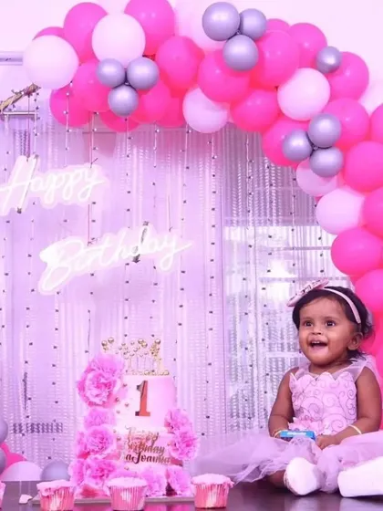 Capturing a precious moment as the birthday girl enjoys her special day. This first birthday setup features a pink and white balloon arch, a shimmering backdrop with a 'Happy Birthday' neon sign, and a custom-designed cake.