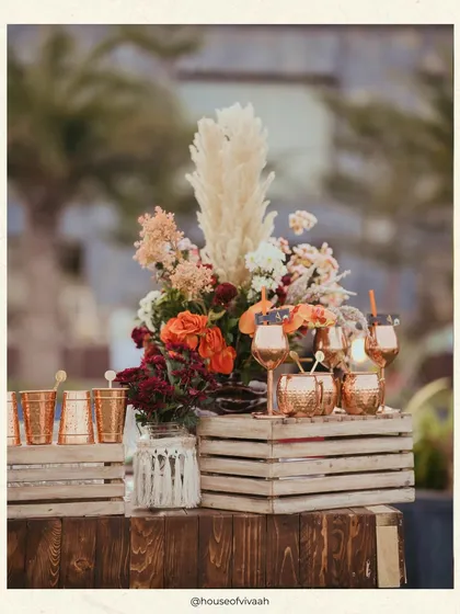 A rustic bar setup with copper mugs and a floral arrangement of pampas and crimson blooms on wooden crates, perfectly capturing the "Mystic Mehendi" vibe.