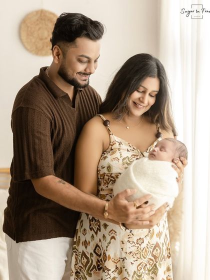 A full-length portrait of a new family in our boho studio. The parents stand together, lovingly holding their swaddled newborn.