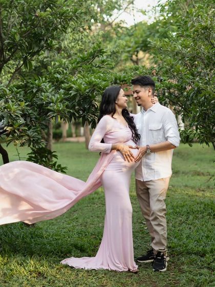 A classic couple's pose in a beautiful park setting. The flowing pink gown adds a pop of color and movement against the green background, creating a romantic and timeless maternity photo.