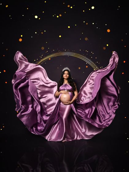 A dramatic solo portrait featuring a flowing purple satin gown against a dark background with golden sparkles. The fabric is tossed to create a beautiful, wing-like shape.