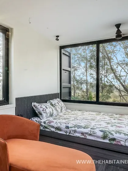A sun-filled bedroom in a container home, with large windows offering views of the surrounding trees.