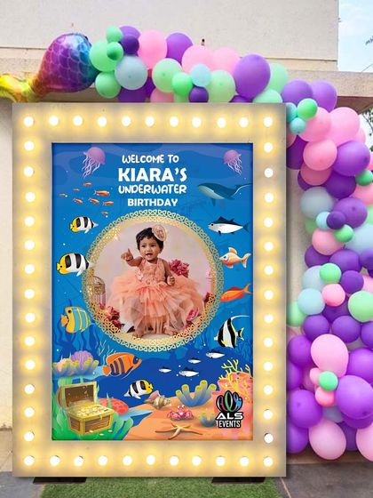 A personalized welcome board for Kiara's underwater-themed party. The board features a photo of the birthday girl and is framed with lights and a balloon garland, making it a great photo spot for guests.