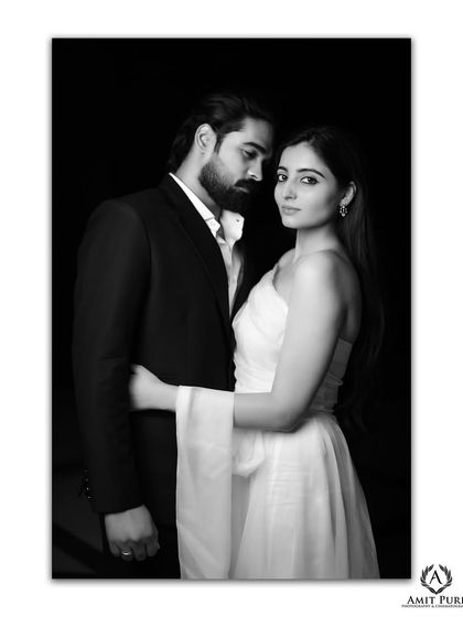 A dramatic black and white portrait against a dark background. The contrast and lighting create a powerful and moody image, emphasizing the couple's striking features and intense connection.
