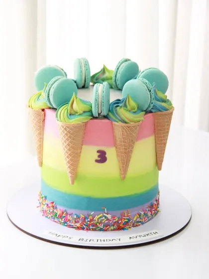 A fun and colorful cake for a 3rd birthday, combining two favorites: ice cream and macarons! The rainbow striped cake is decorated with upside-down cones and a crown of macarons.