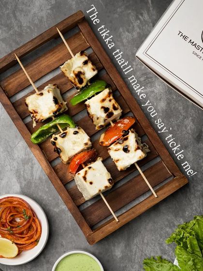 A playful presentation of our Malai Paneer Tikka skewers, arranged on a wooden tray. The creamy white marinade contrasts beautifully with the colorful grilled vegetables.