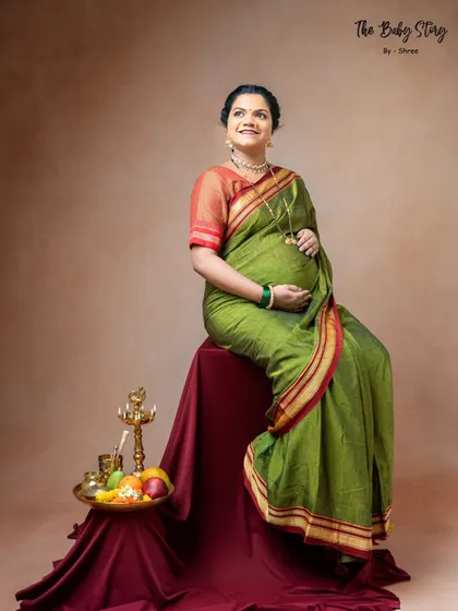 A classic seated portrait from a traditional Maharashtrian maternity session. The mom-to-be looks radiant in a green saree, surrounded by symbolic items like a samai lamp and a platter of fruits.