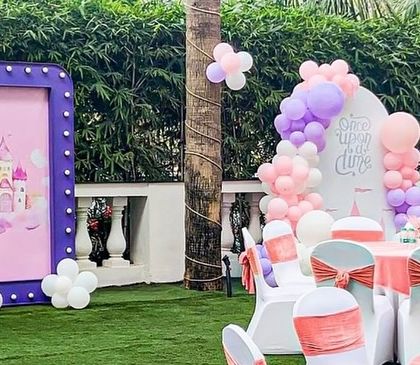 A beautiful outdoor princess party scene. A marquee-lit sign with a castle illustration is paired with a pastel balloon backdrop, creating a perfect photo spot on the lawn.