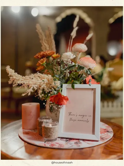 A close-up of a table centerpiece with dried pampas grass, fresh flowers, and a small framed sign that reads "There's magic in these moments."