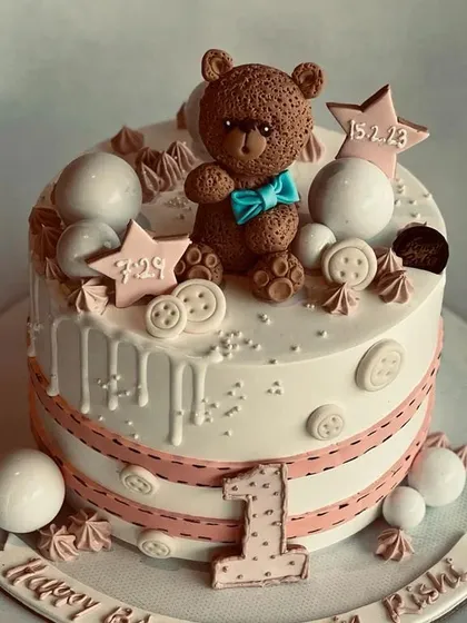 A lovely first birthday cake with a hand-sculpted teddy bear topper. The design includes delicate details like edible buttons, stars, and the baby's birth date for a personal touch.