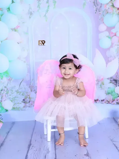 A little girl dressed as a fairy sits on a chair, surrounded by a magical pastel-colored setup. The fairy wings, teddy bears, and floral decorations create an enchanting atmosphere.