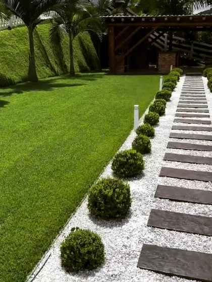 A garden path made of dark stone slabs set in a bed of white gravel, running alongside a lush lawn. The path is lined with neatly trimmed spherical shrubs, adding a formal, structured element to the landscape.