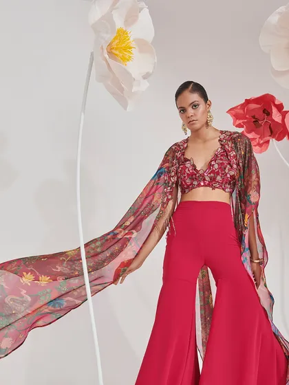 A dynamic shot from a contemporary fashion campaign. The model wears a fuchsia sharara set with a flowing, printed cape, styled to create a sense of movement and vibrant energy against a minimalist floral set.