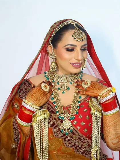 A ravishing bride in a red and gold lehenga, wearing a layered kundan and emerald set with matching kaleeras.