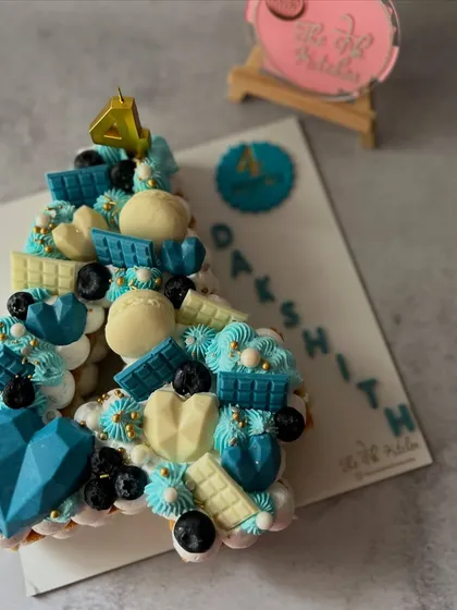 A stylish monogram cake in shades of blue and white for Dakshith's fourth birthday. This number 4 cake is loaded with toppings like blueberries, white and blue chocolate bars, and a geometric chocolate heart.