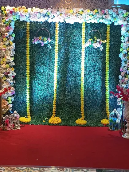 A Haldi backdrop combining a faux green wall with marigold garlands and a floral frame. Decorative lanterns and birdcages on the side add extra detail to the setup.
