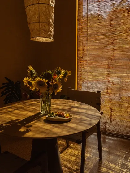 Sunflowers in a vase on a simple wooden table, illuminated by the beautiful, striped light from the bamboo blinds.