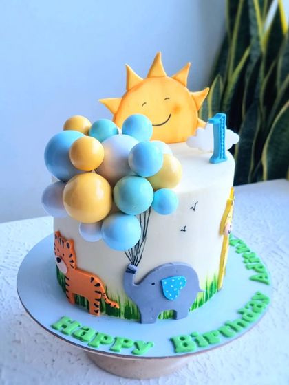 A first birthday cake with a sunny theme, featuring a smiling sun topper and an elephant holding a bunch of balloons. The animals are created with 2D fondant cutouts.