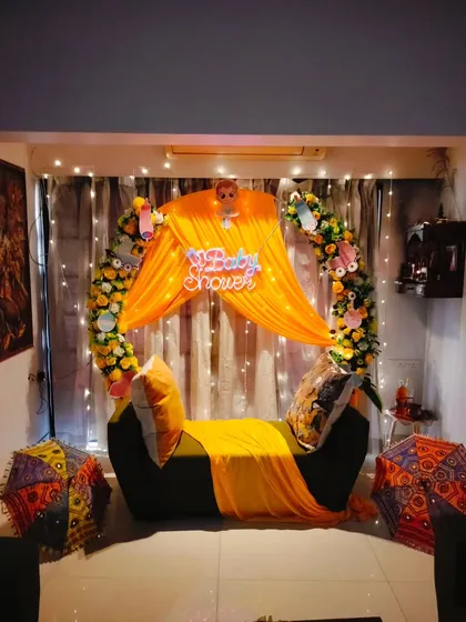A vibrant home baby shower decoration, featuring a sofa decorated with bright yellow drapes, a floral arch, and a "Baby Shower" neon sign.