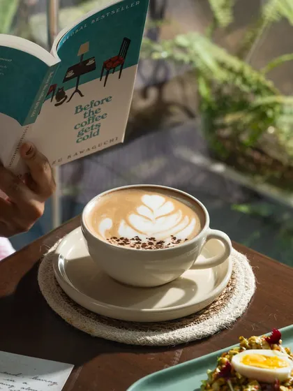 A quiet moment with a book and a cappuccino. This lifestyle shot sells the relaxing atmosphere of the cafe, not just the coffee.