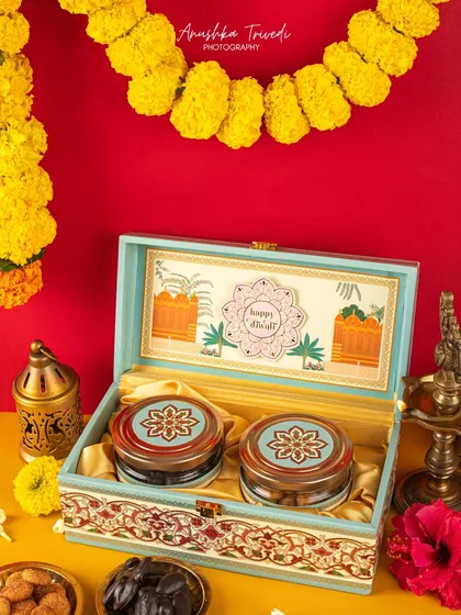 A beautifully crafted wooden box with traditional motifs, filled with jars of dates and cookies. The marigold garland and red background give it a classic Diwali look.