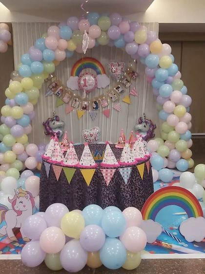 A magical unicorn and rainbow themed party setup. We used a full arch of pastel-colored balloons to frame the cake table, which is decorated with unicorn figures, themed party hats, and a colorful banner.