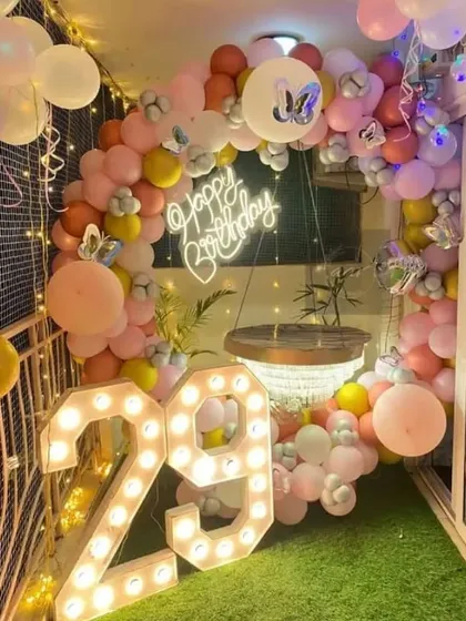 A beautiful balcony decoration for a 29th birthday. This cozy setup features a pastel balloon arch, a neon sign, and light-up numbers on an artificial grass floor.